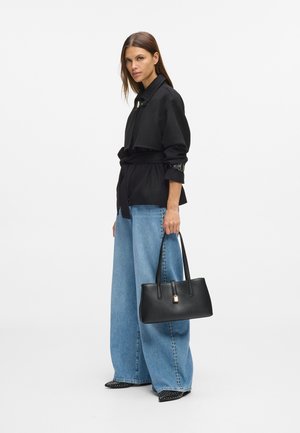 Žena v čiernom sako so zaväzovaním, širokých modrých džínoch so študmi, čiernych špicatých topánkach, drží čiernu kabelku so zlatým zámkom, na bielom pozadí.