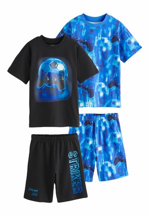 Személy fekete rövidnadrágot visel kék felirattal, amelyen az áll: „Game over In the Zone Level”, beltéren áll egy csíkos párnákkal megpakolt kanapé közelében.