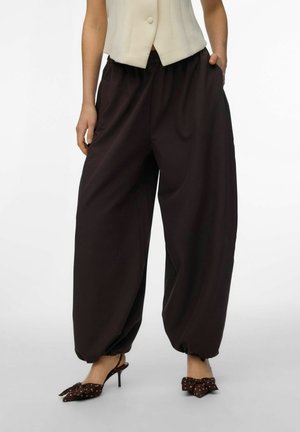 Kobieta ubrana w luźne czarne spodnie z elastycznymi mankietami, beżową bezrękawnikową bluzkę na guziki oraz czarne szpilki z kokardkami w kropki.