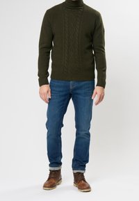 Pull en maille vert foncé à col montant avec un motif torsadé texturé, associé à un jean en denim bleu et des bottes marron à lacets.