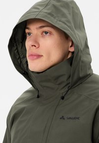 Groene waterdichte jas met een grote capuchon, frontale rits en verstelbare trekkoorden. Bevat een subtiel zwart logo op de borst.