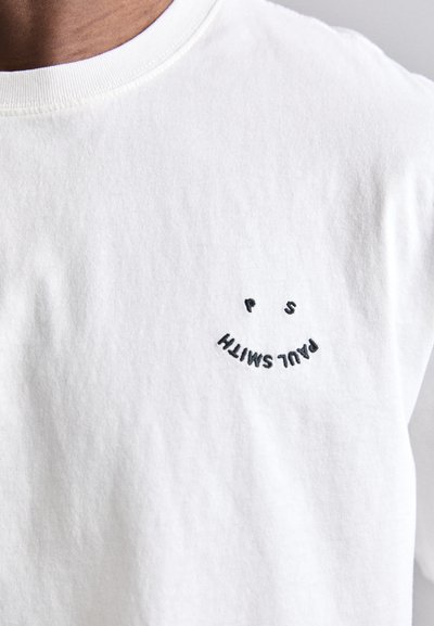 Λευκή βαμβακερή μπλούζα με κοντό μανίκι και στρογγυλή σχεδίαση με μαύρο κεντητό χαμόγελο, καθώς και το κείμενο "PAUL SMITH" σε ημισέληνο κάτω από αυτό.