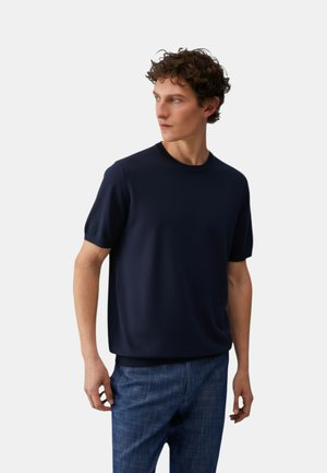 Uomo con capelli ricci che indossa un maglione blu navy a maniche corte e pantaloni blu strutturati, che guarda di lato su uno sfondo semplice.