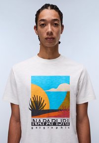 Kremna majica s živahnim grafičkim dizajnom, ki vključuje modro nebo, oranžno sonce in stilizirane rastline; označena z "NAPAPIJRI geographic."