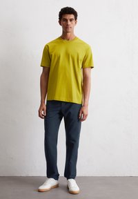 Lime green bawełniana koszulka z krótkim rękawem, w połączeniu z granatowymi spodniami i białymi trampkami z gumową podeszwą, stojąca na jasnym tle.