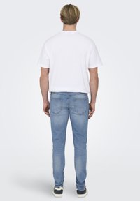 T-shirt bianco in cotone e jeans di denim azzurro chiaro. I jeans presentano due tasche posteriori con dettagli di cucitura. Il modello indossa sneakers casual.