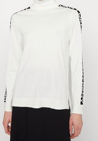 Vit polotröja med långa ärmar. Har svart logotypdetalj längs sidorna. Mjukt stickat material, avslappnad passform, höftlång design.