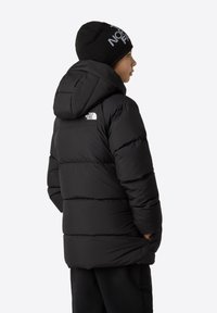 Chaqueta acolchada negra con capucha y costuras horizontales visibles, combinada con un gorro de punto negro que presenta un logo. Textura suave, diseño casual.