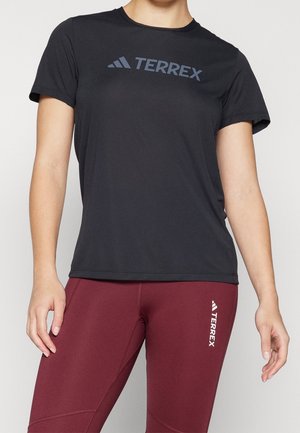 Osoba ubrana w czarną koszulkę Terrex i bordowe legginsy z logo Terrex na udzie, stojąca na jednolitym tle.