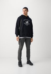 Black hoodie with white logo, paired with grey cargo pants featuring pockets. Worn with black shoes on a plain background.