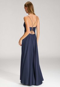 Frau in einem marineblauen bodenlangen Kleid mit dünnen Trägern und einem offenen, geschnürten Rücken, stehend vor einem schlichten, hellen Hintergrund.