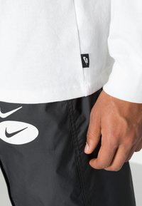 Valge pika varrukaga särk musta Nike sildiga, koos mustade lühikestega, millel on suur valge Nike logo, näidates siledat kangast ja struktureeritud sobivust.