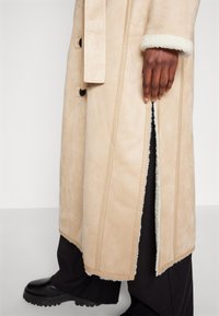 Main à la peau foncée à côté d'un long manteau beige en peau lainée avec une fente latérale, pantalon noir et chaussures noires épaisses sur fond blanc.