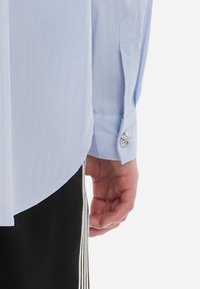 Heledate sinine triibuline pluus, millel on veidi ümar hem, ühe nööbiga mansett ja dekoratiivne nööbi aksent manseti peal.