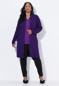 Frau in einem langen lila Cardigan über einem lila Oberteil mit Reißverschluss, schwarzen Hosen, schwarzen spitzen High Heels mit Schleifen und hält eine silberne Clutch.