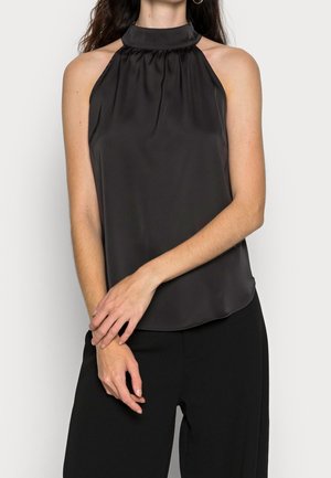 Femme portant un haut noir sans manches en satin avec un col froncé et un pantalon noir, debout devant un fond clair uni.