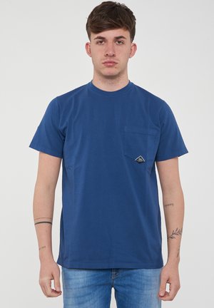 Giovane uomo con capelli castani corti che indossa una maglietta blu semplice con taschino sul petto e jeans blu, in piedi davanti a uno sfondo bianco.