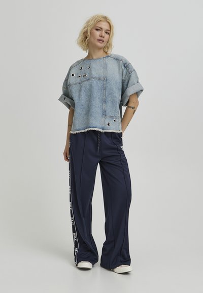 Femme debout portant un haut en denim bleu clair avec des œillets métalliques et des manches retroussées, un pantalon large bleu marine avec des bandes latérales, et des baskets blanches.
