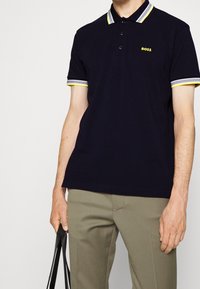 Polo shirt marine avec un col et des poignets rayés jaunes et gris, doté de trois boutons et d'un petit logo "BOSS" jaune sur la poitrine.