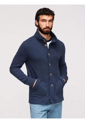 Mężczyzna z brodą, ubrany w granatową zapinaną kurtkę z wysokim kołnierzem i jasnoniebieskie dżinsy, stoi z rękami w kieszeniach na jednolitym tle.