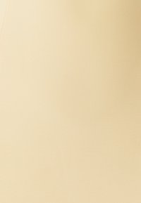 Žena v světle žlutých kalhotách s předními kapsami a stříbrným zapínáním, stojící na jednobarevném bílém pozadí.