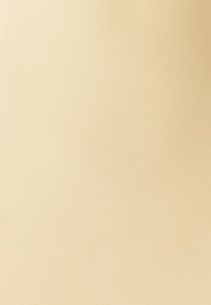 Žena v světle žlutých kalhotách s předními kapsami a stříbrným zapínáním, stojící na jednobarevném bílém pozadí.