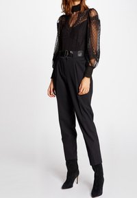 Blusa nera trasparente con pizzo testurizzato e motivi a pois, abbinata a pantaloni neri a vita alta e stivaletti neri con tacco a spillo.