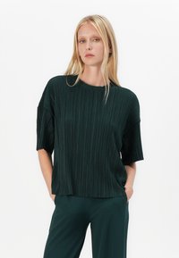 Tamnozeleni, plisirani gornji dio s kratkim rukavima i okruglim izrezom, s opuštenim krojem i mekim materijalom, uparen s odgovarajućim hlačama.