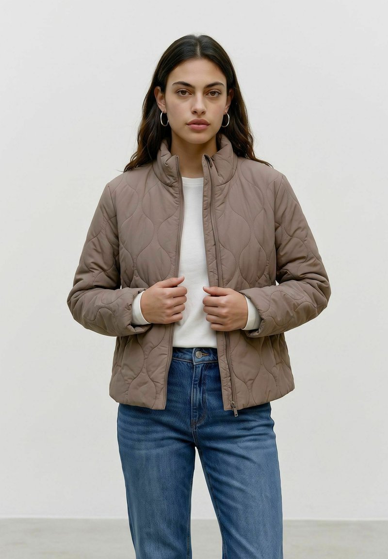 Femme aux cheveux foncés portant des boucles d'oreilles en créoles, une veste matelassée marron clair sur une chemise blanche et un jean bleu, debout devant un fond clair uni.