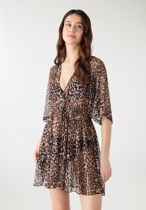 Giovane donna con capelli lunghi e ricci che indossa un abito trasparente marrone con stampa leopardata e vita aderente, in piedi davanti a uno sfondo bianco semplice.