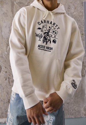 Osoba ubrana w kremową bluzę z kapturem z czarnym, w stylu western, graficznym logo i napisem Carhartt, w połączeniu z niebieskimi dżinsami z białymi wzorami.