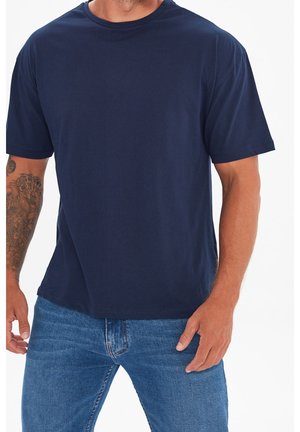 Hombre con camiseta de manga corta azul marino lisa y vaqueros azules, con tatuaje en el antebrazo izquierdo, de pie contra un fondo blanco.