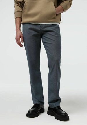 Mężczyzna ubrany w szare spodnie o prostym kroju, czarne masywne buty i beżową bluzę z kapturem, z jedną ręką w kieszeni, na jasnym tle.