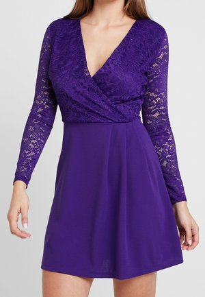 Kobieta w fioletowej sukience z kwiecistą koronką na długich rękawach, dekoltem w serek, dopasowaną talią i rozkloszowanym dołem.