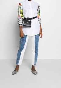 Witte overhemden met kleurrijke, gestreepte mouwen, gecombineerd met blauw-witte color-blocked jeans. Zwarte gestreepte schoenen en een heuptas.