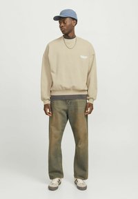 Beige crewneck sweatshirt with graphic, gray ribbed hem, paired with faded blue denim jeans and a blue cap. White sneakers complete the look.