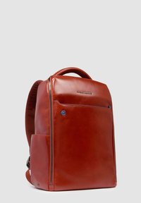 Mochila de cuero marrón con textura suave, bolsillo exterior con cremallera y correas acolchadas. Presenta un logo en plata y acentos en azul.