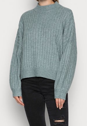 Torso femminile che indossa un maglione a coste grigio chiaro a maniche lunghe e jeans skinny neri strappati.