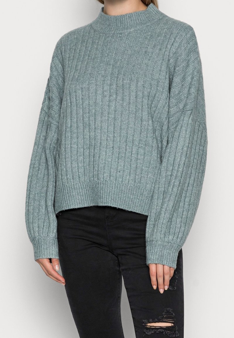 Torso femminile che indossa un maglione a coste grigio chiaro a maniche lunghe e jeans skinny neri strappati.