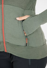 Olivgrüne Zip-Jacke mit strukturiertem Finish, taillierter Passform und kontrastierendem orangefarbenem Reißverschluss. Verfügt über eine seitliche Tasche mit Reißverschluss.