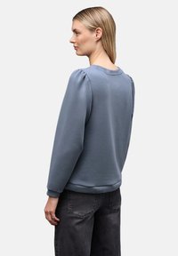 Nainen, jolla on suorat vaaleat hiukset, pukeutuneena siniseen pitkähihaiseen huppariin ja mustiin farkkuihin, seisoo sivuttain tasaisen taustan edessä.