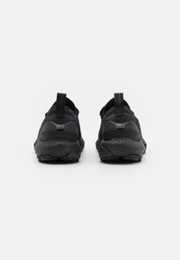 Un par de zapatillas deportivas negras con suelas texturizadas, marcadas como "Phantom" y "HOVR," vistas desde la parte trasera sobre un fondo blanco.