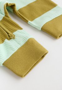 Primer plano de mangas de punto acanalado con bloques de color verde y oliva y un pequeño detalle de cremallera cerca del puño.