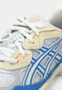 Λευκό, μπεζ και μπλε αθλητικό παπούτσι με διχτυωτό ύφασμα, λευκή κορδόνια και διακοσμητική ραφή στο πλάι και το λογότυπο στη γλώσσα.