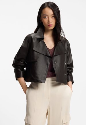 Kvinde iført en sort kort læderjakke over en bordeaux strikket top og højtaljede cremefarvede cargobukser, stående med hænderne i lommerne.