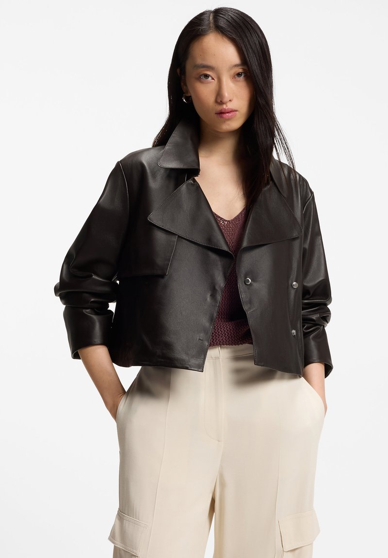 Γυναίκα που φοράει μαύρο κοντό δερμάτινο μπουφάν πάνω από μπορντό πλεκτό τοπ και κρεμ cargo παντελόνι με ψηλή μέση, στέκεται με τα χέρια στις τσέπες.