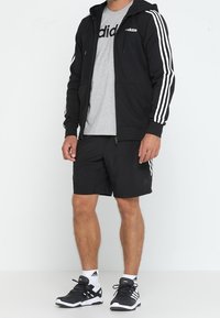 Hombre con chaqueta negra de Adidas, pantalones cortos negros, camiseta gris de Adidas, calcetines blancos de Adidas y zapatillas negras de Adidas, de pie frente a un fondo blanco.