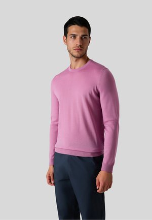 Mand iført en tætsiddende lyserød langærmet sweater og mørkeblå bukser, stående mod en ensartet lys grå baggrund.