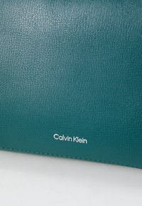 Turkizna denimo denarnica z teksturirano površino, ki na dnu vsebuje belo blagovno znamko "Calvin Klein" in kontrastne šive.