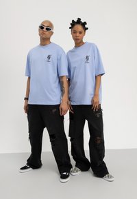 To personer iført lyseblå "Stay Chill" T-skjorter og revne svarte jeans, stående side om side mot en ensfarget bakgrunn.